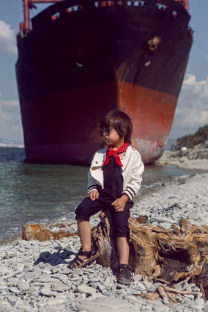 fashionable boy child with long hair sits on a log next to a large ship that ran aground near Novorossiyskの写真素材