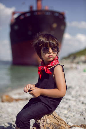 fashionable boy child with long hair sit on a log next to a large ship that ran aground near Novorossiyskの写真素材