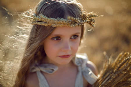 girl child in a dress and a wreath on her head stands on a mown field of wheat at sunset in summerの写真素材