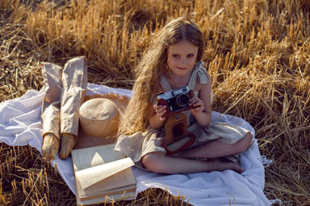 girl child in a dress sitting on a mown field with a camera on a blanket with bread and a book
