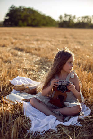girl child in a dress sitting on a mown field with a camera on a blanket with bread and a bookの写真素材