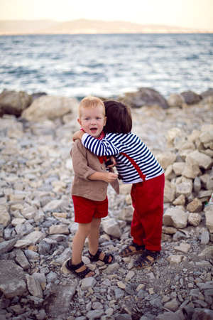 two stylish baby boys in vests stand on the seashore of pebbles in summerの写真素材