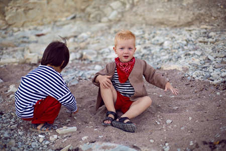 two stylish baby boys in vests sit on the seashore of pebbles in summerの写真素材