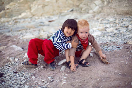 two stylish baby boys in vests sit on the seashore of pebbles in summerの写真素材