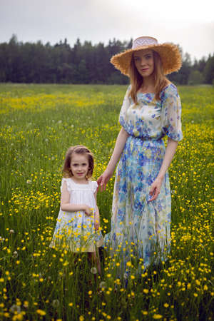 mother and daughter in dresses and a hat stand in a field of yellow flowers in the summer dayの写真素材
