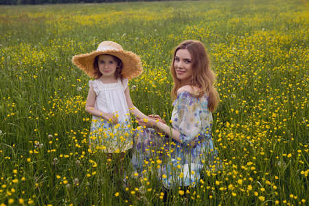 mother and daughter in dresses and a hat stand in a field of yellow flowers in the summer dayの写真素材