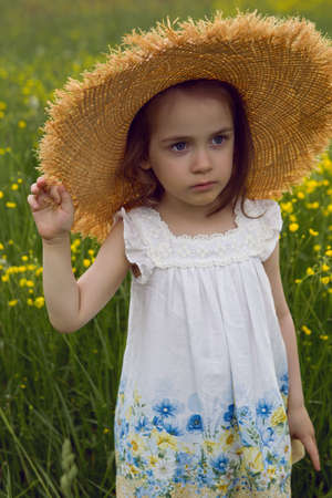 child girl in a straw hat and a dress with flowers stands on a yellow fieldの写真素材