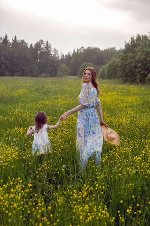 mother and daughter in dresses and a hat stand in a field of yellow flowers in the summer dayの写真素材