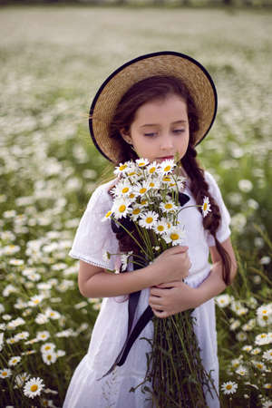 portrait girl child in a white dress stands on a camomile field in a hat and with a bouquet of flowersの写真素材