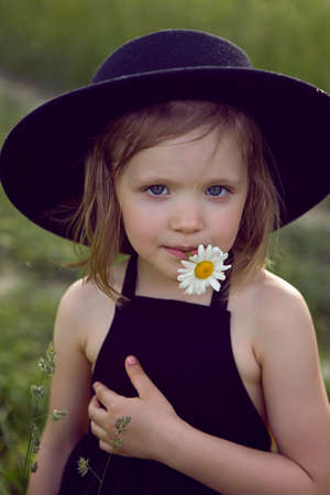 child girl in a black hat and dress stands in a field with a camomile in her mouth in summerの写真素材