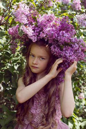 Caucasian girl child seven years old in a purple dress stands in nature with a wreath of lilacsの写真素材