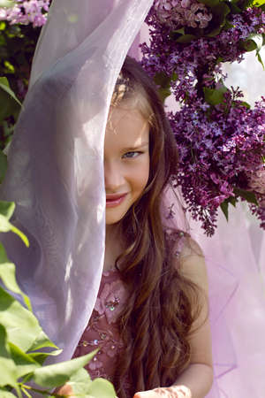 Caucasian girl child seven years old in a purple dress stands in nature with a wreath of lilacsの写真素材