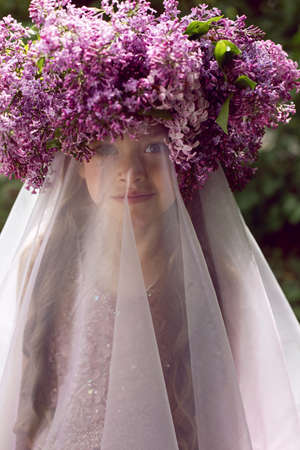 Caucasian girl child seven years old in a purple dress stands in nature with a wreath of lilacsの写真素材