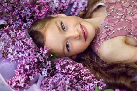 child girl in a purple floral dress lies on the ground among lilacs on a veil in springの写真素材