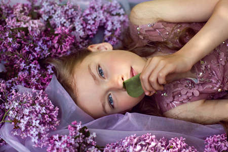 child girl in a purple floral dress lies on the ground among lilacs on a veil in springの写真素材