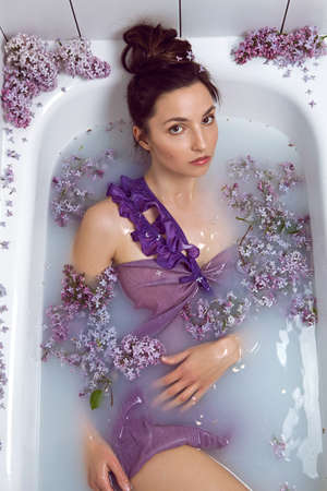 sexy fashionable woman in swimsuit dress lies in a white bath with milk and lilac flowersの写真素材