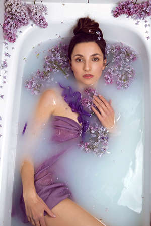 sexy fashionable woman in swimsuit dress lies in a white bath with milk and lilac flowersの写真素材