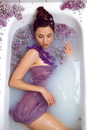 sexy fashionable woman in swimsuit dress lies in a white bath with milk and lilac flowersの写真素材