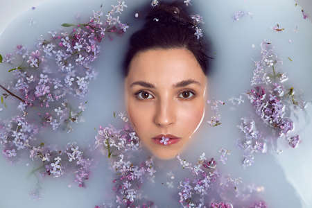 fashionable woman lies in a white bath with milk and lilac flowers