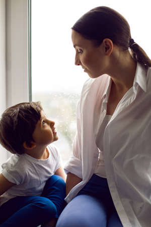 mom and son are sitting in a white shirt and a T-shirt on the window of a house in spring and having funの写真素材