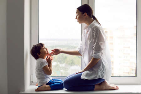 mom and son are sitting in a white shirt and a T-shirt on the window of a house in spring and having funの写真素材
