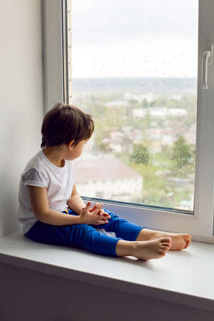boy in a white tank top and blue pants sits on a window in rainy weatherの写真素材