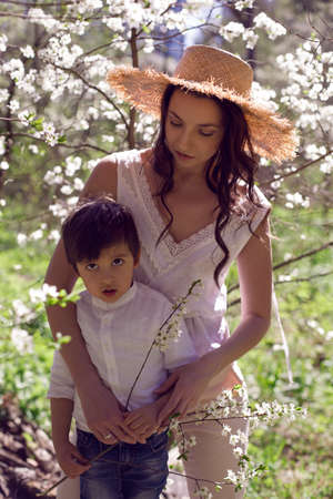woman in pink clothes and a straw hat stands with a child in a white shirt in a blossoming apple orchardの写真素材