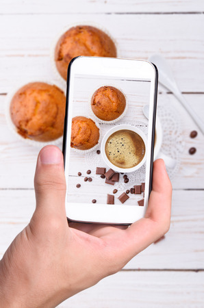 Hands taking photo cup of coffee with muffin with smartphone.の写真素材