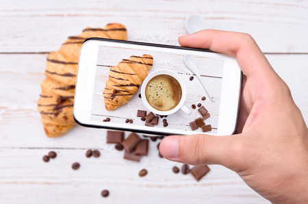 Hands taking photo cup of coffee with croissants with smartphone.の写真素材
