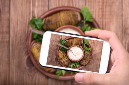 Hands taking photo baked potatoes with sauce on plate with smartphone.