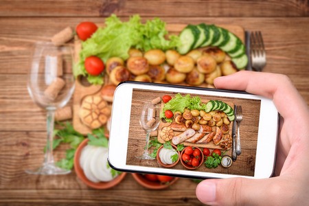Hands taking photo grilled sausages and vegetables on the wooden board with smartphone.