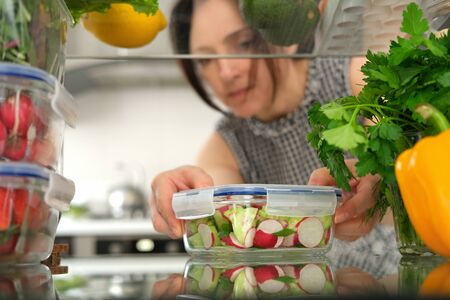 Woman looking inside a fridge full of food and choosing a salad in the container.