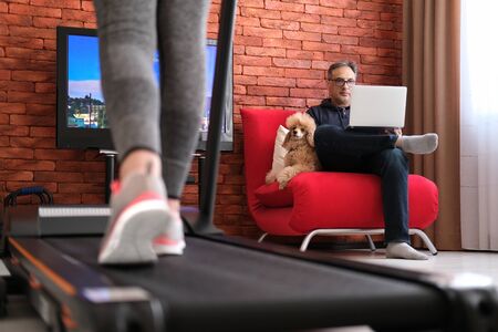 The man is working remotely while woman exercising on a treadmill at home.の写真素材