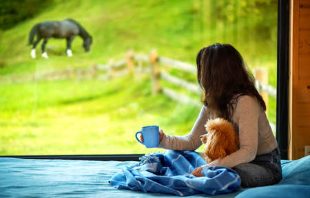 Woman sitting with her dog on the bed and looks outside the window seeing a mountain.の写真素材