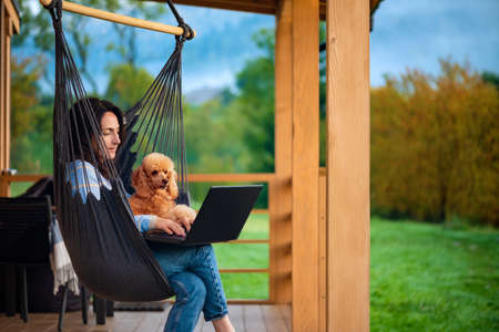 Young freelancer woman with dog working at the computer on the hanging chair on the terrace. Concept of the workplace at home, working remotely.の写真素材