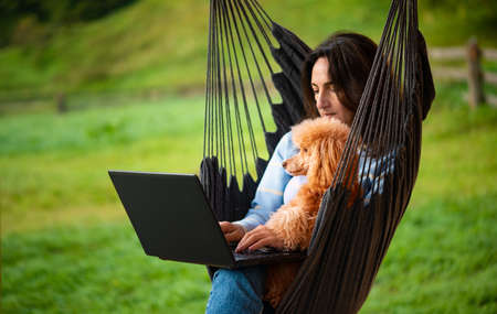 Young freelancer woman with dog working at the computer on the hanging chair on the terrace. Concept of the workplace at home, working remotely.の写真素材