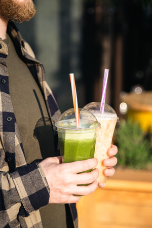 Man outdoors holding plastic cups with green and orange smoothies, close up.