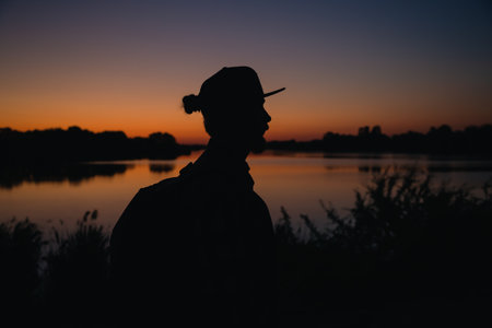 A bearded man with backpack and with cap at sunset near the lake.の写真素材