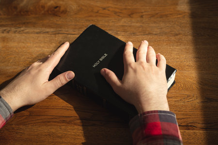 Hands of a Christian man on a Bible book on a wooden table.