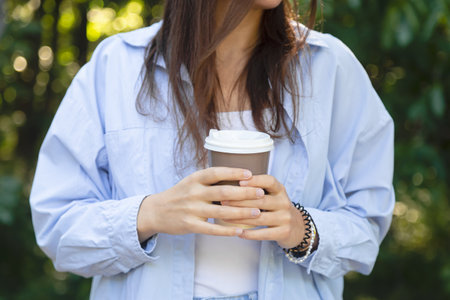 A young woman holds a cup with coffee in her hands on a background of nature.