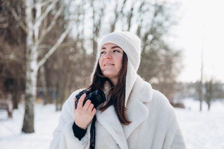 A young woman in a fur coat and hat with a camera in a winter park.の写真素材