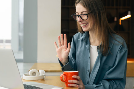 A young woman with glasses sits in front of a laptop and waves hello.の写真素材
