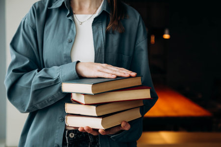 A woman in a blue shirt holds a stack of books on a dark library background.の写真素材