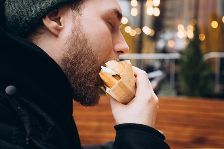A young bearded man eats a burger outside against the backdrop of a cafe.の写真素材