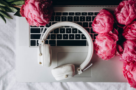 White headphones, laptop and pink peonies on white sheets in bed, top view.