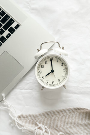 White alarm clock and laptop on a white bed, top view.の写真素材