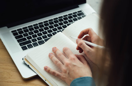 A woman takes notes in a notepad using a laptop sitting at the table.の写真素材