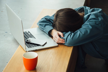 A tired woman lies face down in front of a laptop with a cup of tea.の写真素材