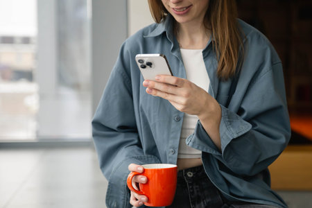 A woman is using a smartphone and holding a cup while sitting by the window.の写真素材