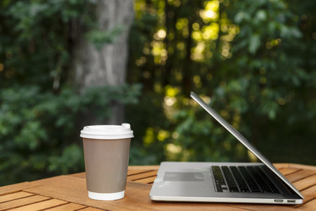 A paper cup of coffee and a laptop on a wooden table on a blurred forest background, work in nature, on vacation and remotely, copy space.の写真素材